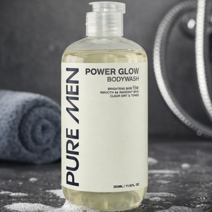 Bottle of Pure Men Power Glow Bodywash on a dark surface with a towel and shower head in the background.